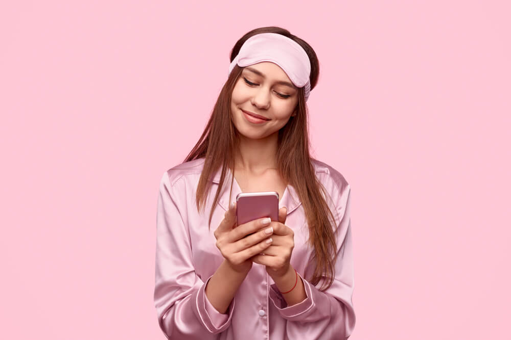 Cute teen girl in pajama and sleep mask smiling and browsing smartphone before going to bed against pink background.
