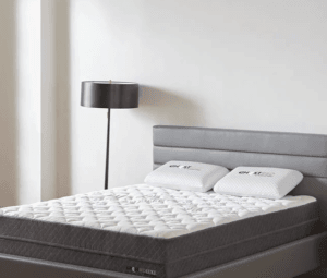 A white mattress with two white bed pillows on a gray fabric bed frame