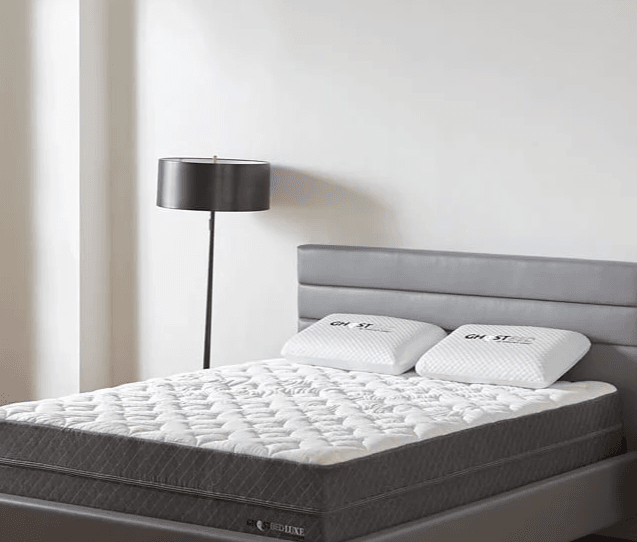 A white mattress with two white bed pillows on a gray fabric bed frame