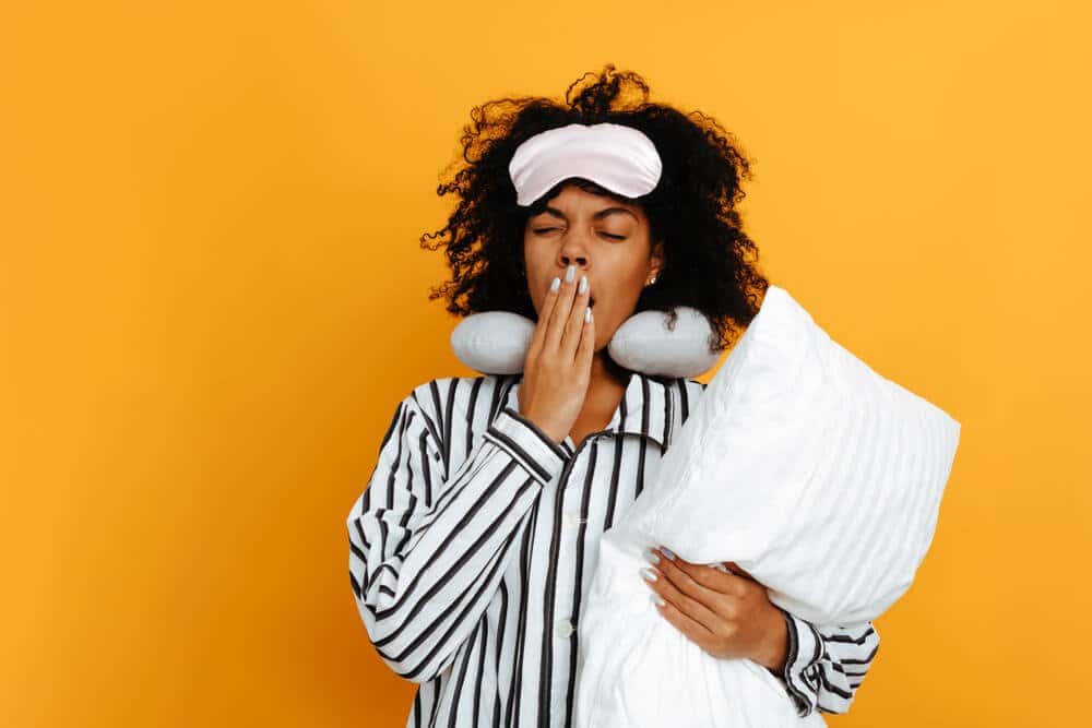 Sleeping. Dreams. Woman portrait. Afro American girl in pajama and sleep mask is holding a pillow and yawning, on a yellow background.