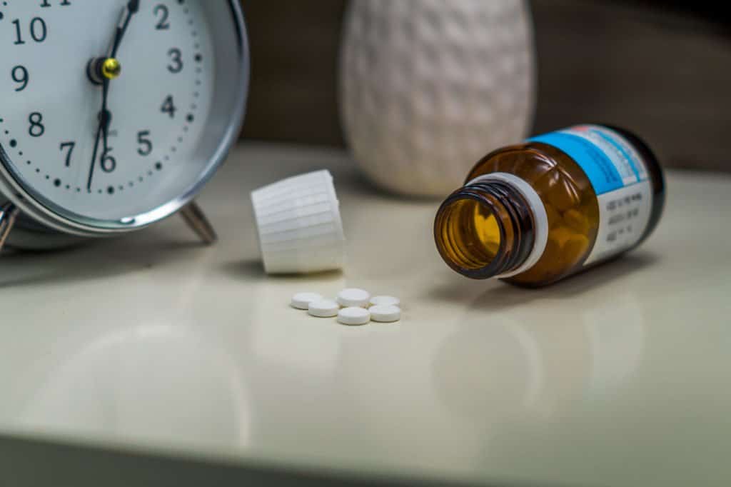 A brown pill bottle spilling white pills on a white nightstand with an alarm clock in the background.