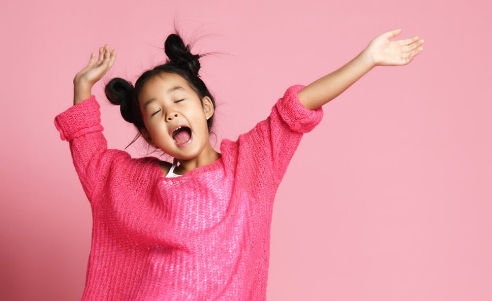 Asian kid girl in pink sweater, white pants and funny buns sings singing dancing on pink background; sleep music for kids concept.