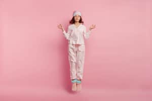 A woman meditating and wearing pink pajamas and standing in front of a pink background.