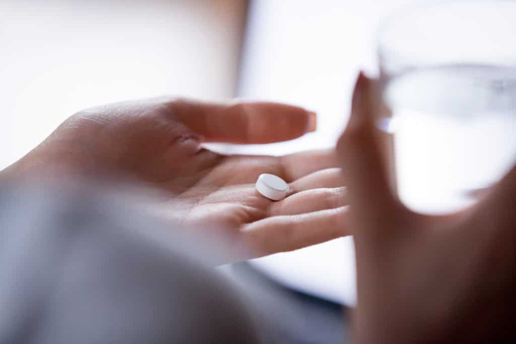A hand holding a white pill and a glass of water.