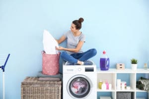 Beautiful young woman with clean laundry sitting on washing machine at home; how to wash bamboo sheets concept.