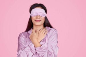 Young lady in sleep mask touching delicate silk sleepwear and enjoying softness and comfort while resting against pink background