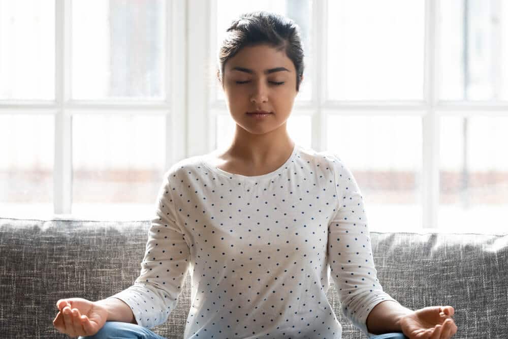 Calm Indian woman with closed eyes meditating at home alone close up, relaxed beautiful girl sitting in lotus pose on comfortable couch, dreaming, doing yoga exercise, stress relief concept .