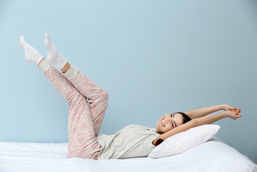 Young woman in pajamas stretching herself on bed over blue background.