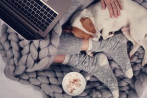 A woman sitting on a wool blanket with her dog, a laptop and a cup of coffee.