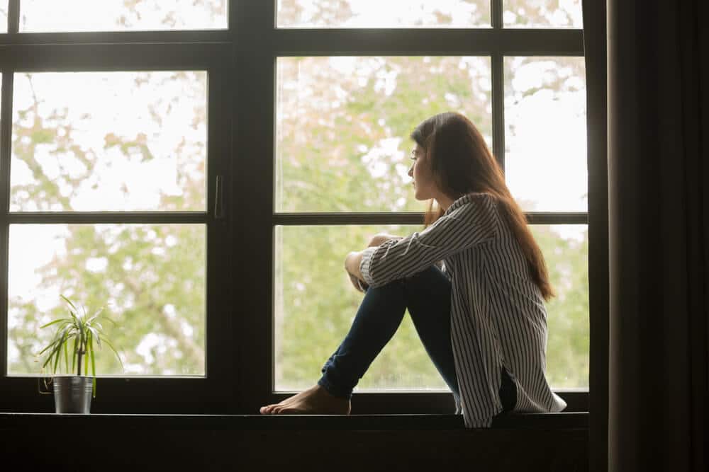 Thoughtful girl sitting on sill embracing knees looking at window, sad depressed teenager spending time alone at home, young upset pensive woman feeling lonely or frustrated thinking about problems with adhd and sleep.