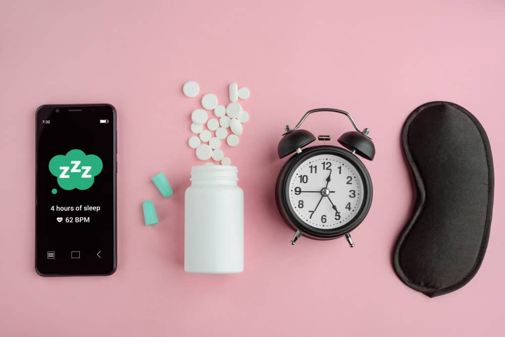 Phone, earplugs, pills, table clock and mask on pink background, flat lay. App tracker, medicine and accessories for good sleep.