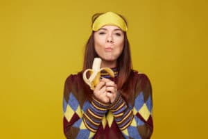 Lovely mixed race young woman holds delicious banana, tastes with pleasure, enjoying likes eating sweets, has good appetite, wears colorful sweater and sleeping mask on head. Healthy eating concept.
