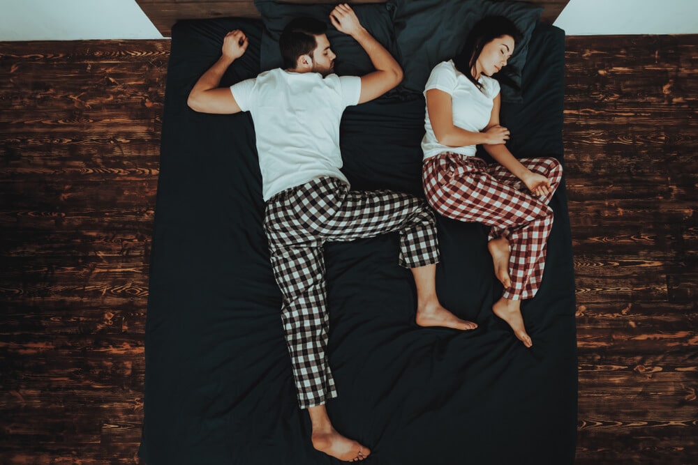 Couple is Lying on Bed. Couple is Young Beautiful Woman and Man. People is Sleeping on Bed. People is Wearing Pajama Pants and T-Shirts. Persons is Located in Home Interior. Top View.