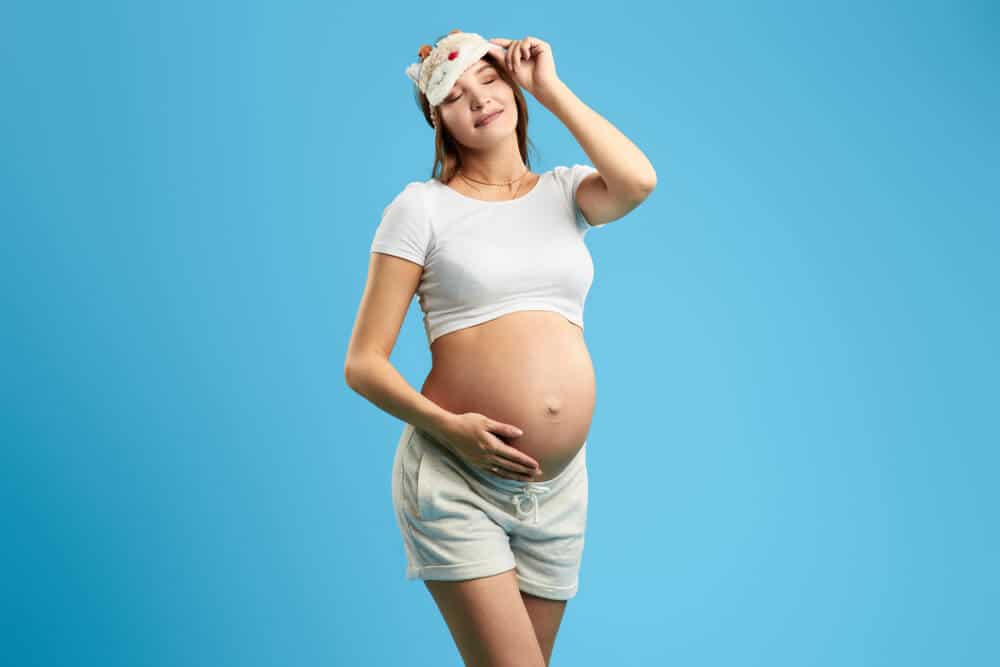 sleepy girl with closed eyes touching her belly, enjoying pregnancy, isolated blue background. studio shot. happiness, love, family, copy space