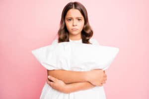 A young girl holding a white pillow and frowning in front of a pink background.