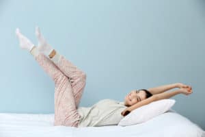 Young woman in pajamas stretching herself on bed over blue background