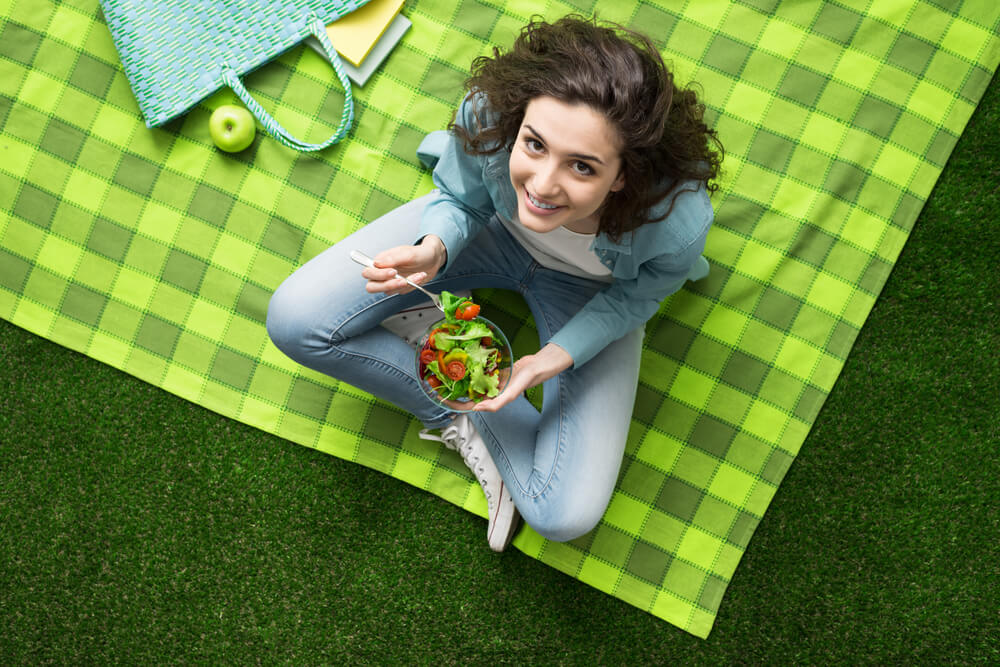 Smiling woman having a relaxing lunch break outdoors, she is sitting on the grass and eating a salad bowl