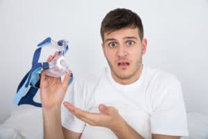 A young man holds his CPAP mask with a confused face.