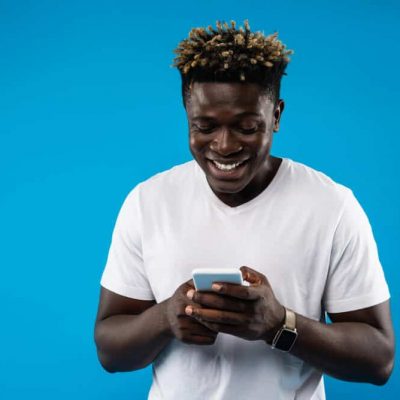 Portrait of young man in white t-shirt using mobile phone and smiling. Isolated on blue background