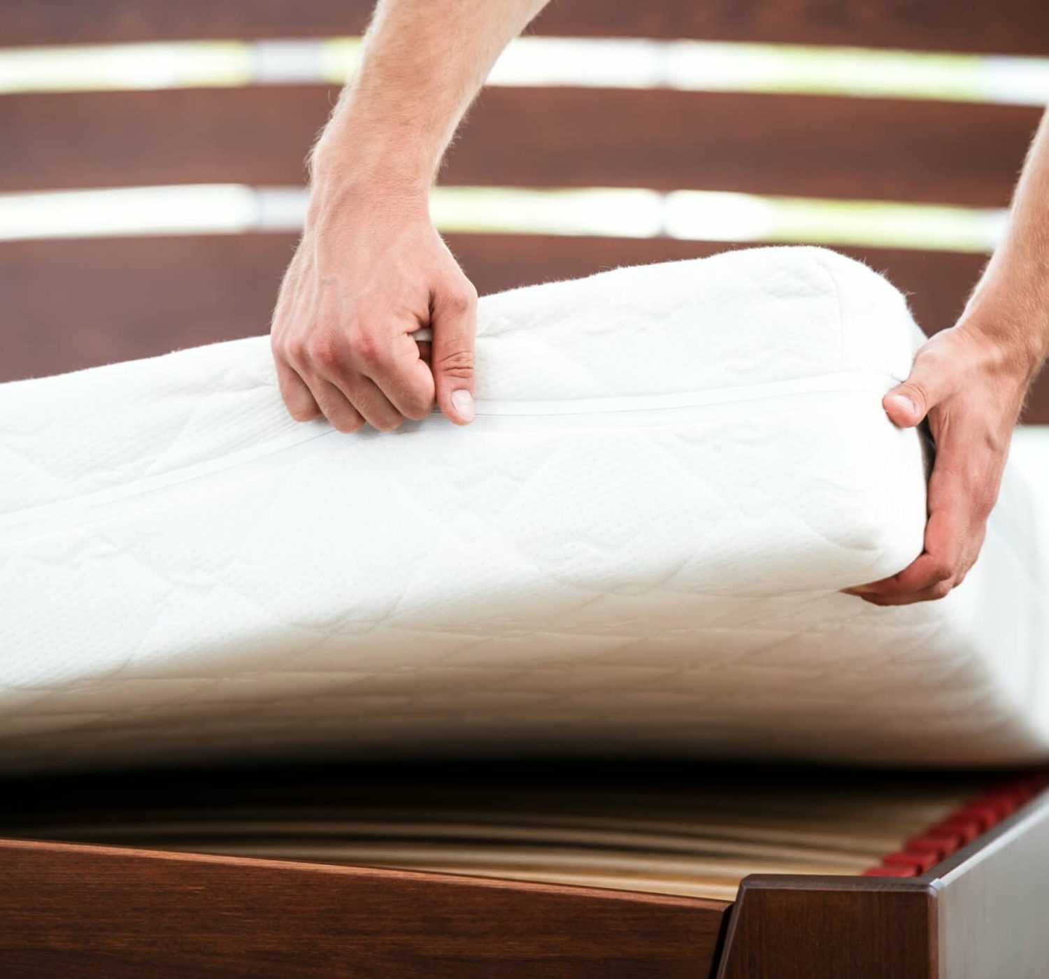 Two hands laying a white mattress on a brown slatted bed frame.